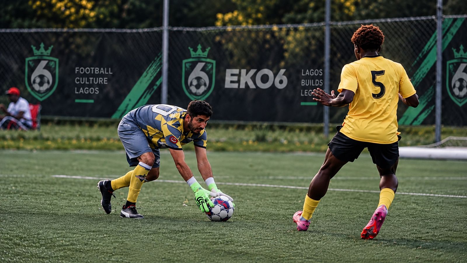 Goalkeeper gathering the ball as an attacker closes in, with EKO6 banners on the fence behind